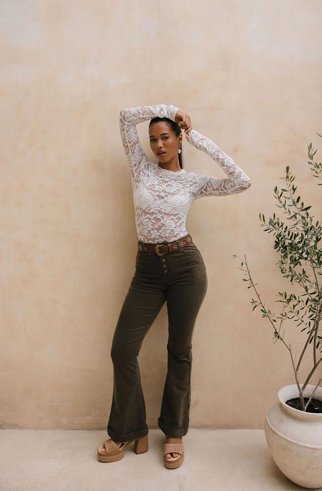 Woman wearing a white lace top and dark pants against a beige wall with a plant.