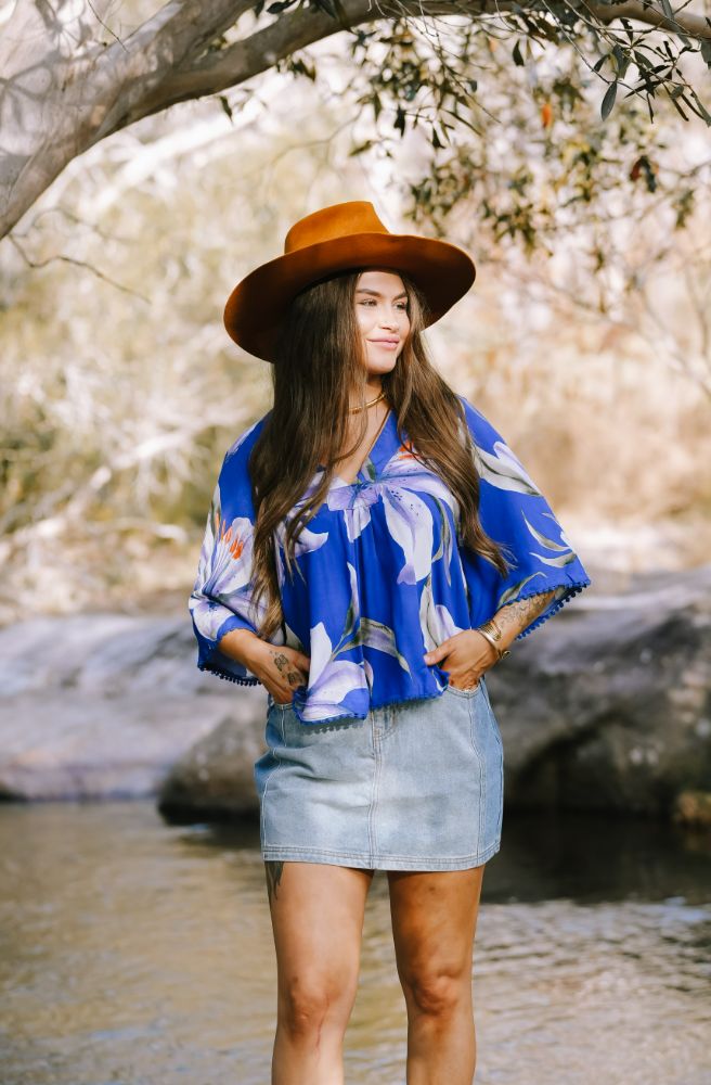 Woman wearing a blue floral blouse and denim skirt standing by a river with trees in the background