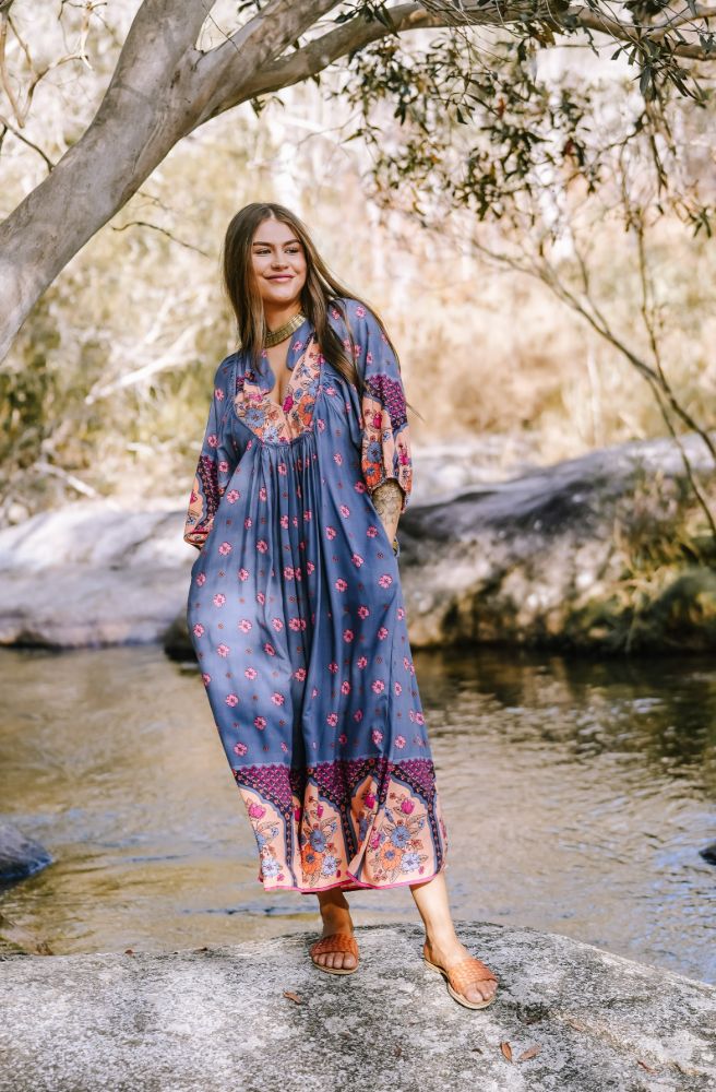 Woman in a long, patterned dress standing by a stream with trees in the background