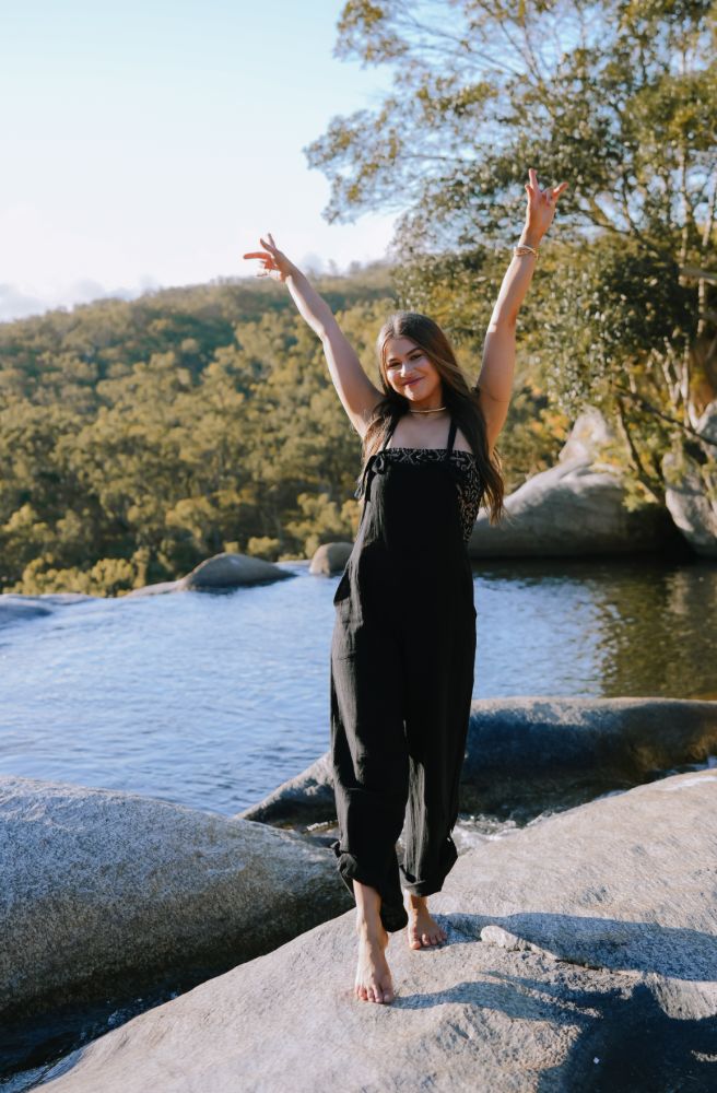 Woman standing on a rock by a body of water with trees in the background with comfy black overalls on