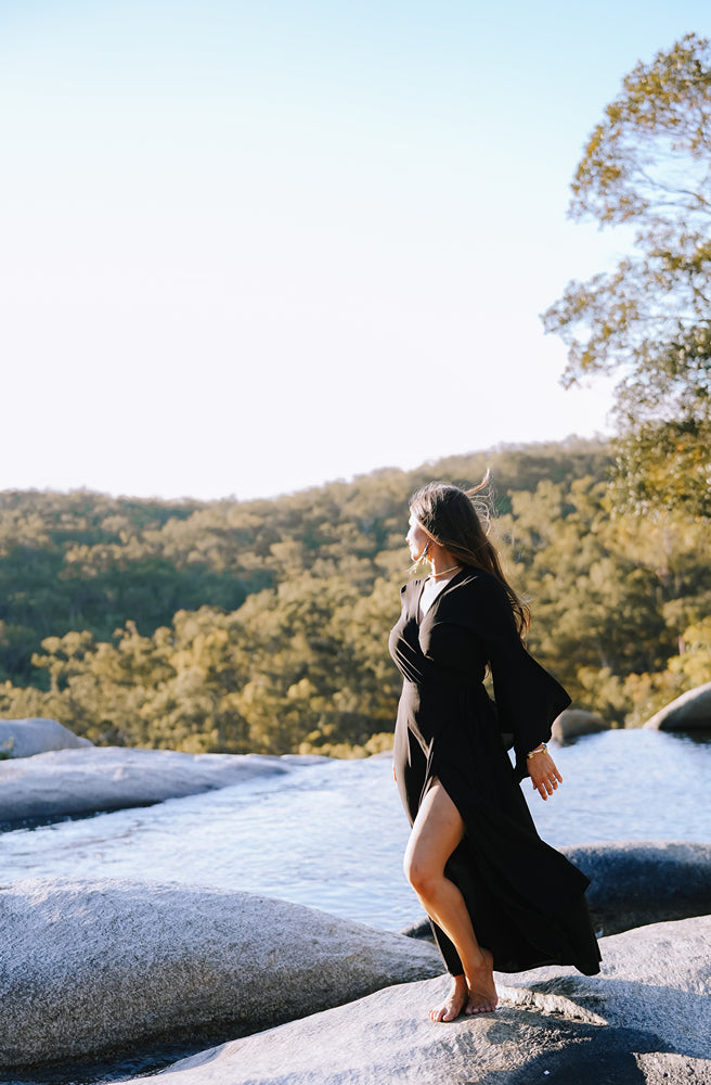 Woman in a black dress standing on rocks with a scenic background of trees and mountains.