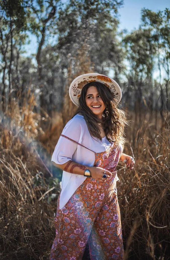 Earth Essence Transit Overalls, A woman standing in a field wearing floral overalls with thin adjustable spaghetti straps, a square neckline, and a hat.