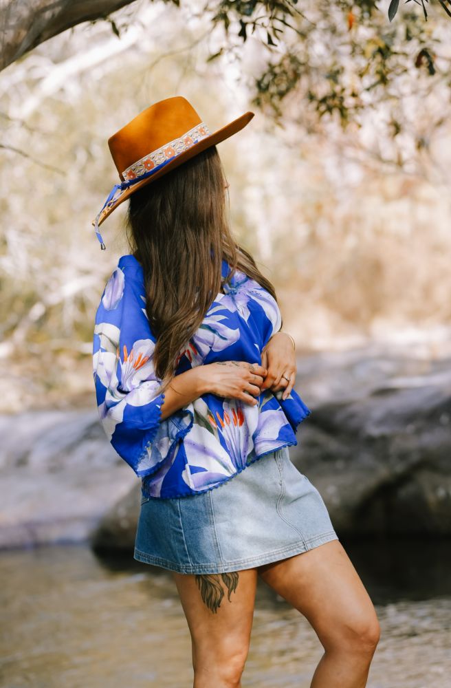 Woman wearing a blue floral shirt and denim skirt with a brown hat, standing by a stream.