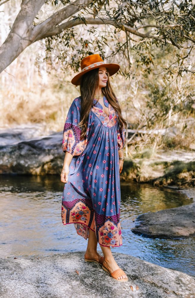 Woman in a colorful dress and hat standing by a stream with trees in the background
