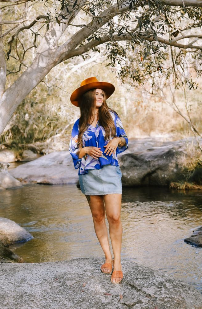 Woman in a blue shirt and hat standing by a stream with trees in the background