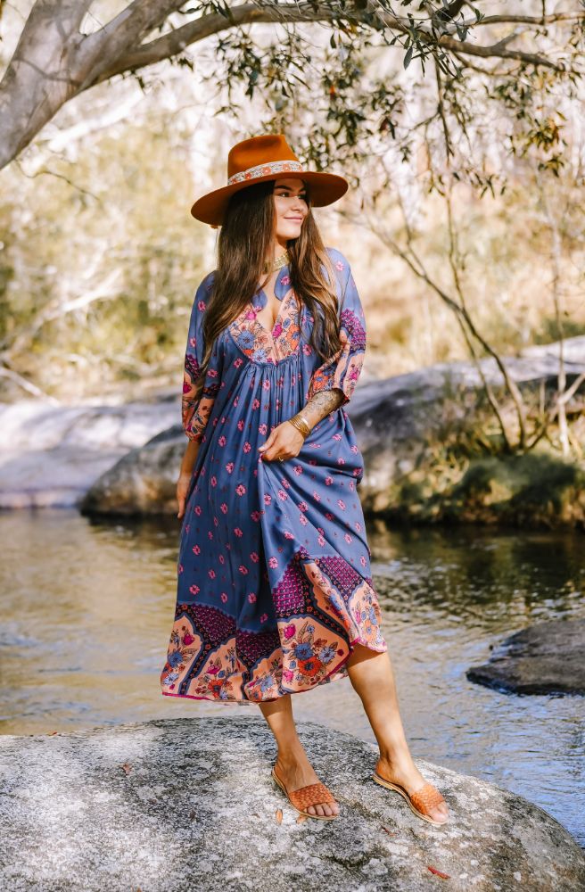 Woman in a blue floral dress and hat standing by a stream