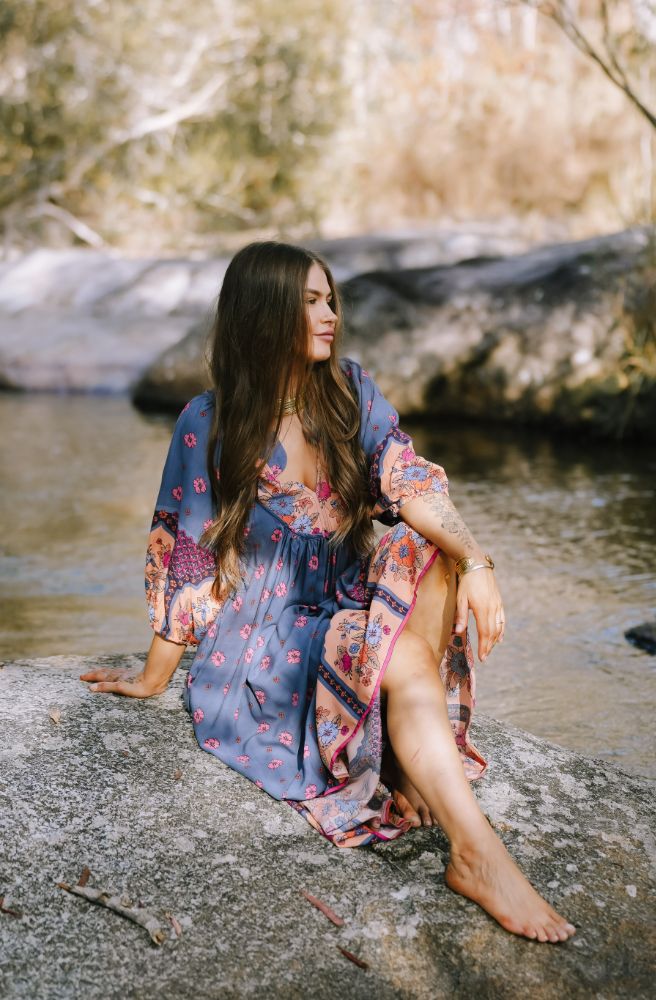 Woman in a colorful dress sitting by a stream with trees in the background floral boho dress