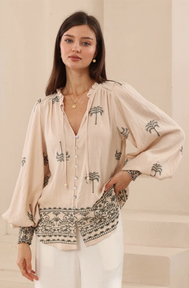 Sahara Palms Blouse Shirred Cuff, Woman wearing a tan blouse with dark green palm tree print and white pants against a beige background