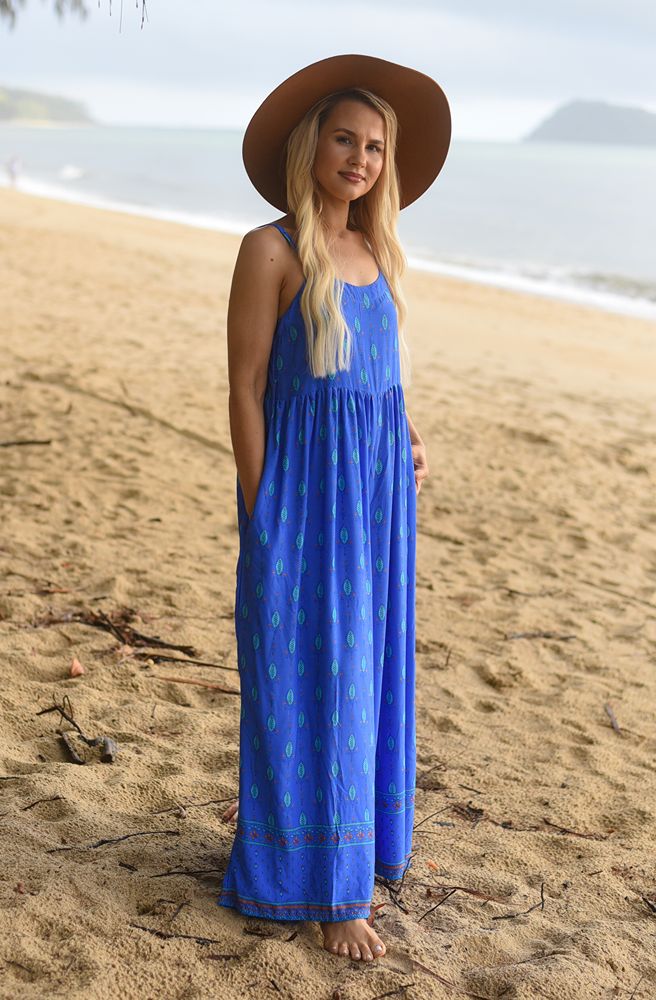 Woman in a blue dress and brown hat standing on a sandy beach.