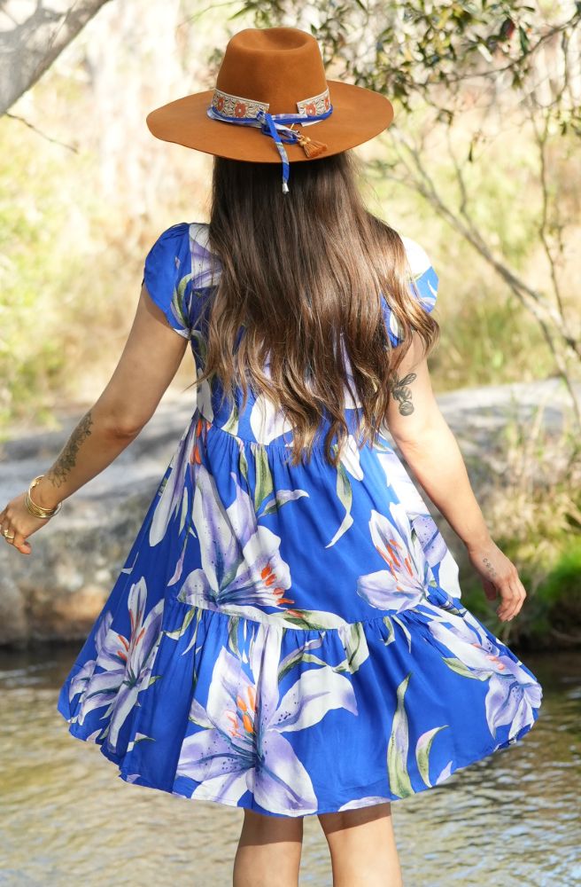 Woman in a blue floral dress and brown hat standing by a water body.