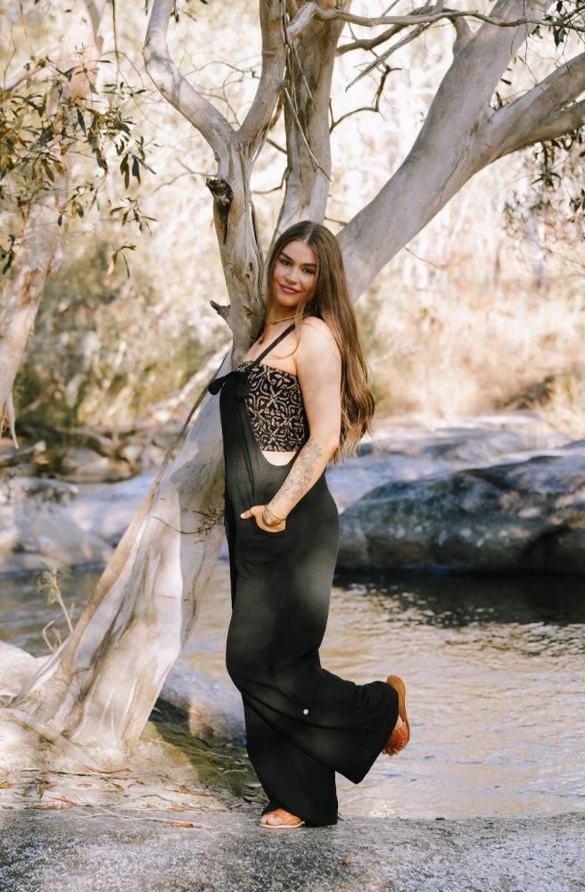 Woman in a black outfit standing by a tree with a natural background black cotton overalls 
