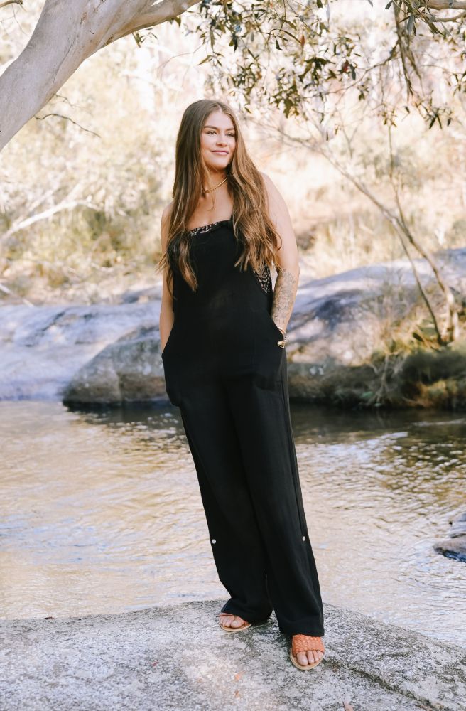 Woman in a black outfit standing by a stream with trees in the background with black cotton overalls on