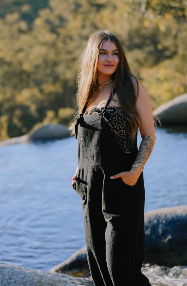 Woman in black overalls with leopard print top standing by a body of water with trees in the background one piece overalls on 