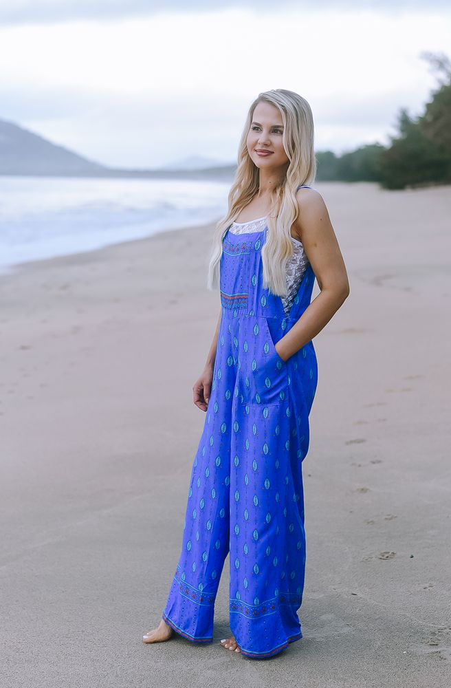 Woman in a blue jumpsuit standing on a beach with ocean and sky in the background