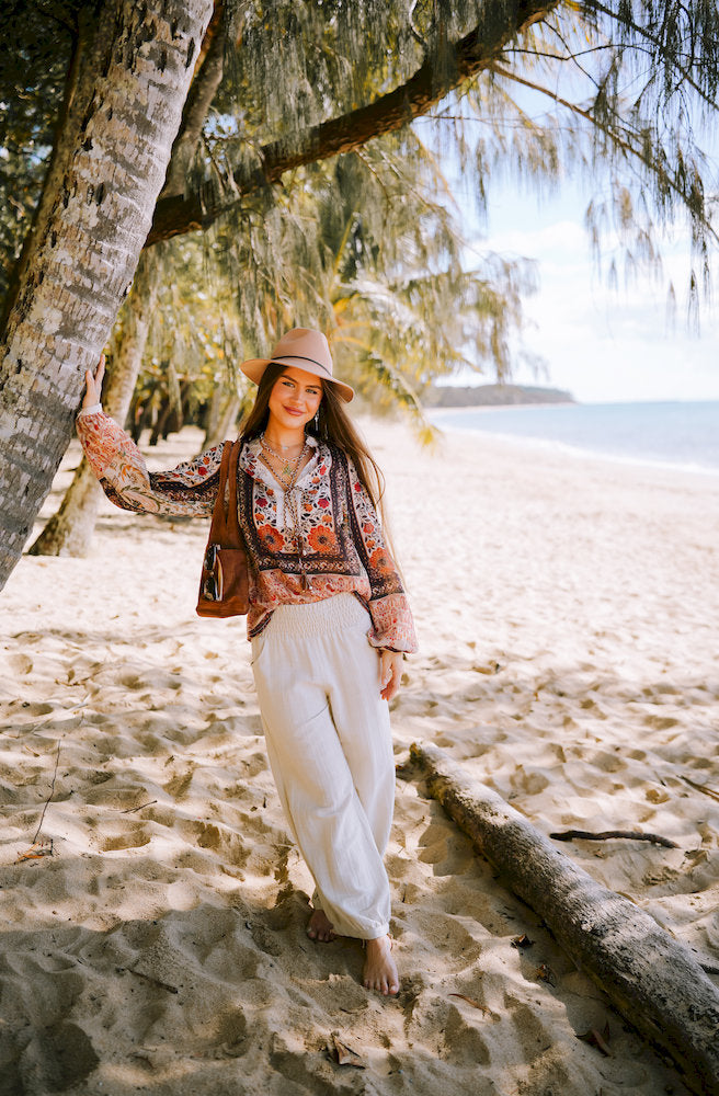 Woman in a floral boho blouse and linen pants leaning on a palm tree at the beach