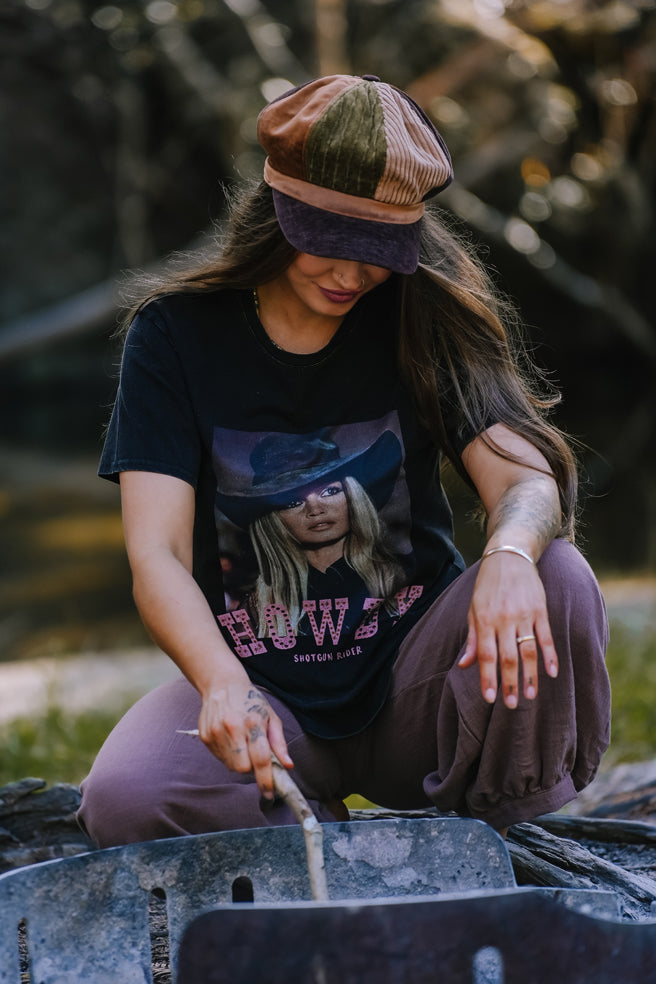 Person sitting outdoors, wearing a cap and t-shirt with a graphic design, near a grill.