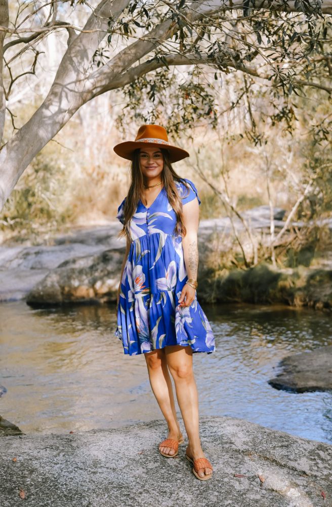 Woman in a blue floral dress and hat standing by a stream with trees in the background