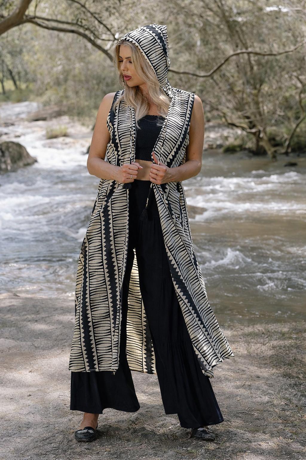 Hemi Hood Kimono, Woman wearing a long black and cream patterned sleeveless kimono with hood over a black outfit by a river.