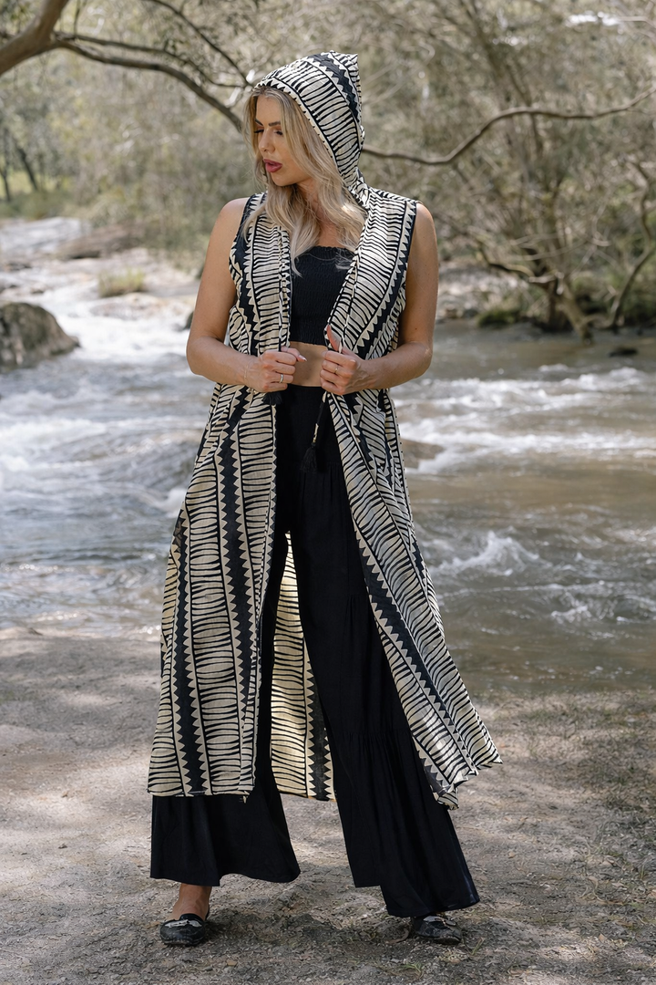 Hemi Hood Kimono, Woman wearing a long black and cream patterned sleeveless kimono with hood over a black outfit by a river.