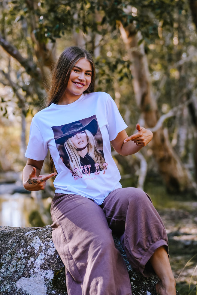 Woman wearing a white t-shirt with a graphic design, sitting outdoors among trees. Camping clothing