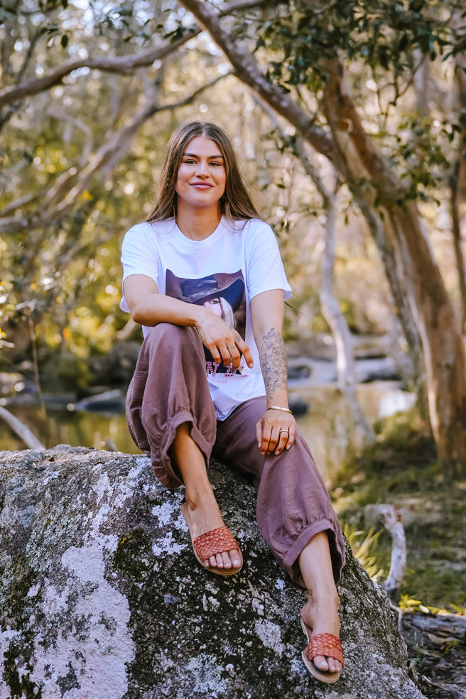 Woman wearing a white t-shirt with a graphic design, sitting outdoors among trees. Camping clothing Far north 