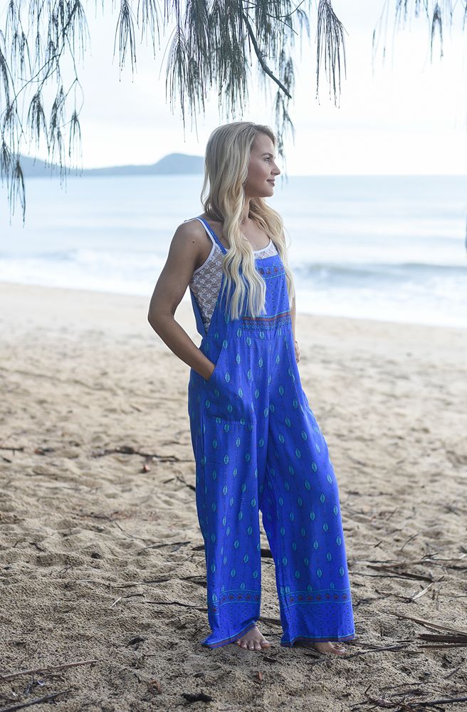 Woman wearing a blue jumpsuit standing on a beach with ocean and trees in the background
