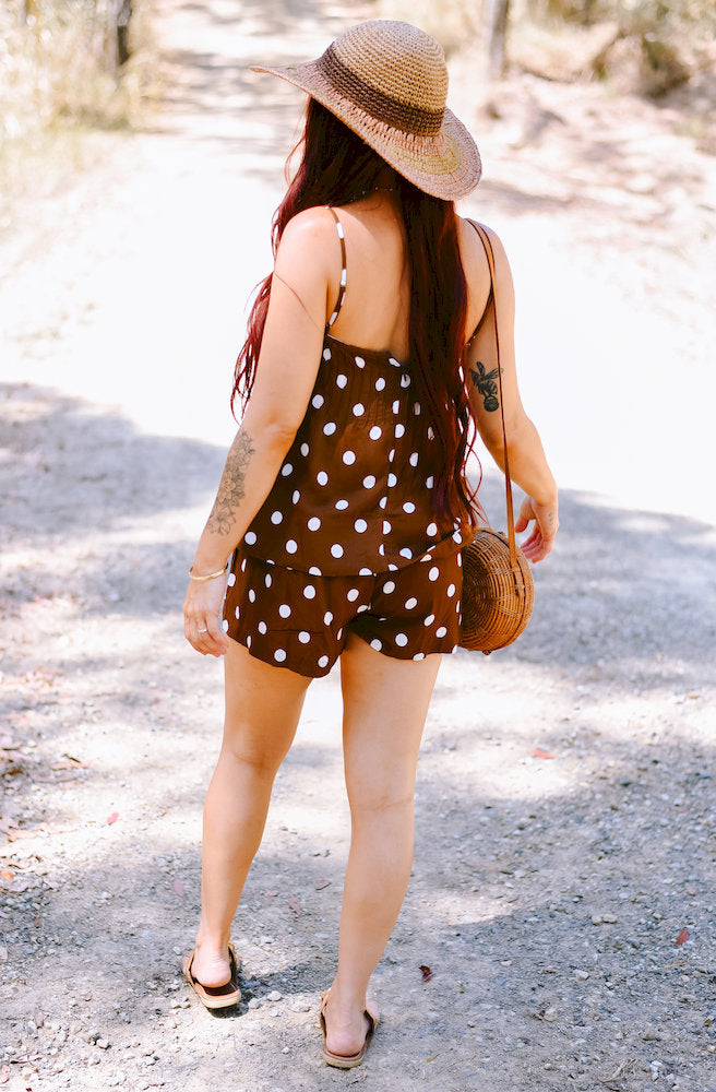 Women's brown playsuit with white polka dot print styled with sandals and a woven bag.